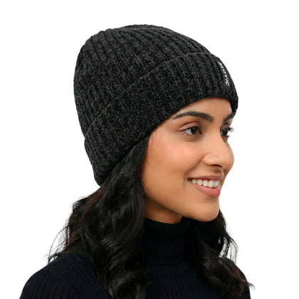 A woman wearing a dark charcoal gray ribbed knit beanie with a small white embroidered logo on the side, photographed against a clean light gray background.