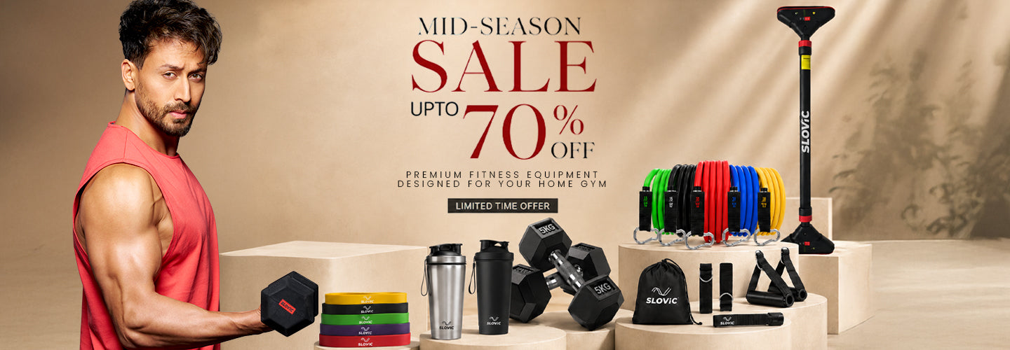 A fitness-themed promotional banner featuring Tiger shroff  in a red sleeveless shirt holding a Slovic dumbbell on the left, with various gym equipment and accessories displayed on pedestals on the right. Large text reads 'MID-SEASON SALE UPTO 70% OFF'.